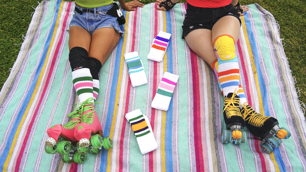 Two skater sitting on a blanket. Photo is focused on the Moxi x Skater Socks they are wearing. There are 4 pairs of Skater Socks laid next to them.
